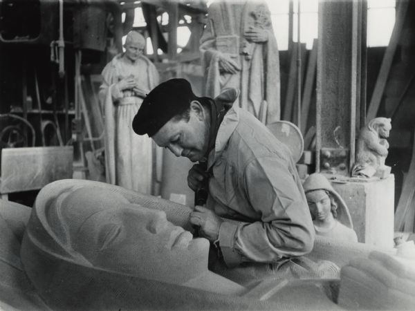 A man is leaning over a large sculpture of a woman and carving it. He is wearing a beret, a scarf, and a workshirt. Several other sculptures — all apparently of religious themes — are behind him.