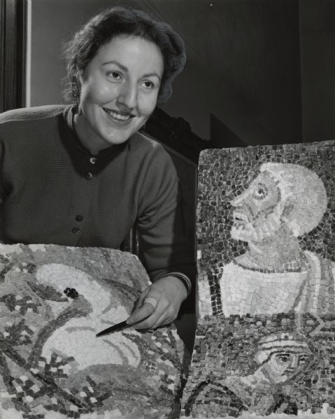 A woman is smiling while pointing to a tile on a mosaic she is displaying in front of her. Two other mosaics are next to her. The caption reads: "A lecture and demonstration on mosaics was given Wednesday night by Miss Mary Basso, 236 W. Wright St., at a meeting of the Studio Club at Milwaukee-Downer College. She showed copies of ancient mosaics she brought home from Italy."