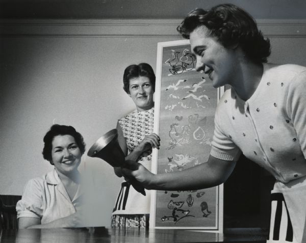 A woman in the foreground is holding a bell, while two other women in the background are smiling at her. The woman on the right is standing and holding a framed work of art. Caption reads: "The ringing of a bell will mean not that dinner is served but that another picture has been sold at the third annual country art fair. The outdoor sale is sponsored by the Friends of Art of the Milwaukee Art Center. Mrs. James Grootemaat, 5168 N. Kent Av., Whitefish Bay, held a serigraph by a Milwaukee artist, Schomer Lichtner. The bell ringer was Mrs. Ernest J. Philipp, Jr., 3934 N. Stowell Av., Shorewood, co-chairman of the fair. Both wore the sturdy little aprons in which the Friends of Art will store money and tickets."