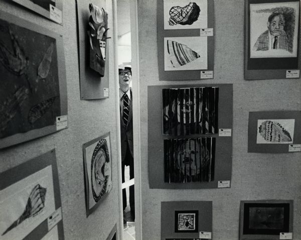 Two room divider panels are displaying several works of art. Framed between the two panels is a man standing on the other side of the panels. Caption reads: "Paul Wisneski, of Milwaukee, looked over an art display Monday at the Grand Avenue retail center. Milwaukee Public School students' artwork will be exhibited through March 20."