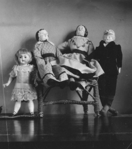 Four dolls are posed together. Two porcelain dolls are seated on a small wicker chair, and the chair is flanked by two other dolls. A caption reads: "Doll at left, 50 years old. Center dolls 75-80. Boy doll 45 yrs."