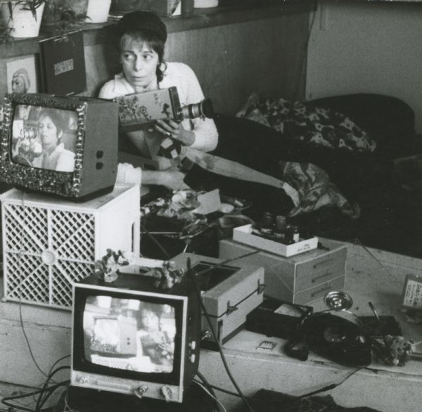 Shirley Clarke is sitting on a couch and holding a video camera. She is looking off to the side. There are two monitors and other equipment in front of her, and her image is on both monitors.