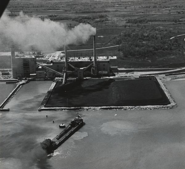 Aerial view of a large ship with a considerable amount of dark smoke coming from the rear. The ship appears to be heading toward a large field of coal built out into the water. Caption reads: "The coal ship J.R. Sensibar churned the muddy water at her stern as she tried to free herself Monday from a mud bank where she went aground Sunday. She had driven her bow into the mud while maneuvering in wind to enter the Electric Co.'s Oak Creek dock. The ship was pulled free by a tug Monday night."