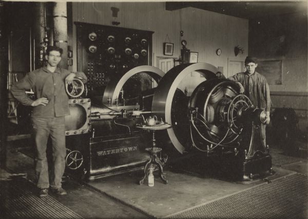 Two men are standing on either side of an engine in a room. Displays with dials are on the wall behind them. A small, round table with a teapot on it is in front of the engine, and a bottle is sitting on the ground beneath it. Caption reads: "Engine room, 1911. Loyal Baxter, 22 years of age, engineer standing at right. Curley Nelson, 22 years of age, fireman."