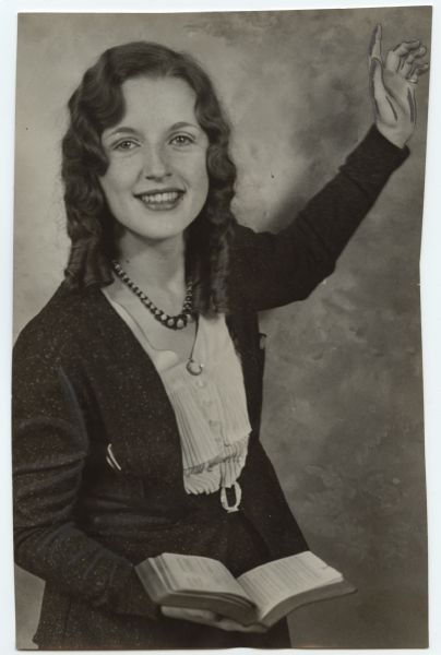 A smiling woman is posing with an open book (probably a Bible) in one hand, and has her other hand raised. Caption reads: "Preaches Here. A girl evangelist from Hollywood, Calif., only 17 but boasting six years experience, is preaching in Milwaukee for two weeks. She is Halleine Smith and is holding her meetings at the Paris theater, Twenty-second and Center Sts., every evening. A few years ago she was expelled from a girls' school in California for daring to call the principal a 'sinner.' Halleine is with her mother and younger brother on a trip east."