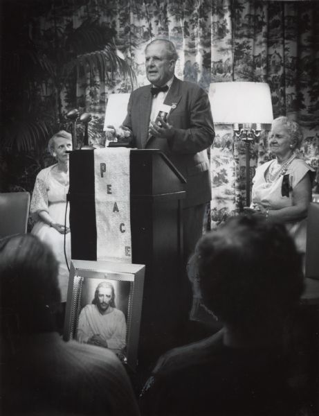 A man wearing a suit and bow tie is standing at a podium, speaking and gesturing with his hands. A lectern scarf is draped over the podium with the word "Peace" on it. A framed painting of Jesus is propped on the floor in front of the podium. Two women are sitting behind the man, and two other people are sitting in the foreground. Caption reads: "A Wisconsin hotel room was dedicated Wednesday night as a sanctuary for the annual convention of the Federation of Spiritual Churches and Associations. The Rev. Bert Welch of Brea, Calif., spoke to delegates. The convention will continue through Sunday." 