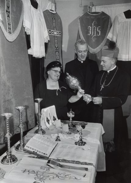 Two men in clerical collars (and one wearing a biretta, cross, cassock, and cincture) examining a monstrance held by a woman. Several clerical vestments are hanging above them, and a table with church paraments, an open Bible, chalices, and altar candlestick holders is next to them. Caption reads: "A variety of religious items made and acquired by the Missionary Association of Catholic Women was examined Tuesday. From left are Mrs. Joseph [Mary] Gockel, 2464 N. 39th St., national president; Father Walter C. Dean, 1409 N. Prospect Av., national spiritual director, and Bishop Harold W. Henry of the diocese at Kwangju [Gwangju], Korea. They participated in the 39th annual convention of the association at St. Rose school, N. 30th and W. Clybourn Sts."