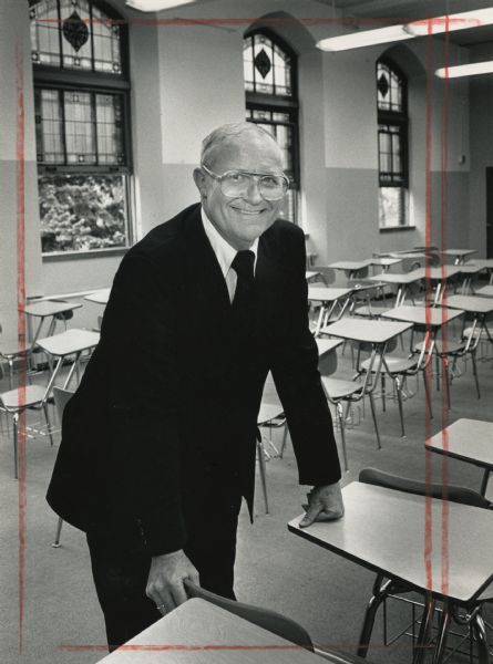 A man is smiling and leaning against a school desk. Caption reads: "Brother Norbert Karpfinger will be leaving Milwaukee." 