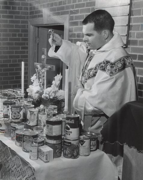 A man wearing an alb and chasuble is blessing a collection of canned goods on an altar. Caption reads: "Canned food collected for the St. John's Home for Aged Women was blessed Thanksgiving day by the Rev. James Hynek, vicar of St. Paul's Episcopal church, 7929 W. Lincoln Av., West Allis. The food was collected by children."