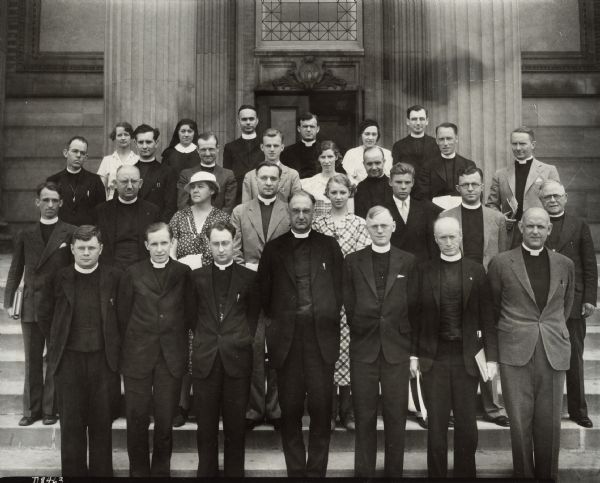 Twenty-nine people, both clergy and laypersons, are posing for a group portrait in front of a campus building.