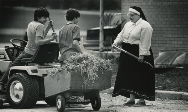 Two boys are sitting and standing next to a four-wheel utility vehicle that is hauling a cart of hay. They are facing a woman standing on the right who is wearing a nun's veil and is holding a shovel. Caption reads: "WORK DAY — Chris and Pat Bohman took a break from their work and stopped with their load to talk to Sister Mildred Rieber during a recent cleanup of the grounds at Holy Trinity Catholic Church in the Village of Newburg."