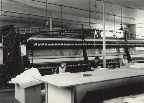 Three women are sitting behind a long table and working with lace. Large machinery is behind them.