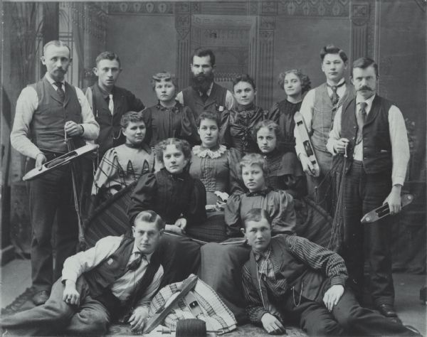 Studio group portrait of fifteen men and women posing together in front of a painted backdrop. Some of the people are holding weaving tools.