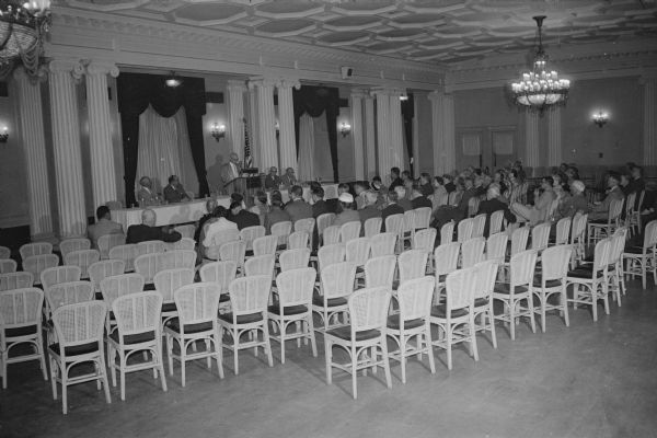 View from back of room towards a group of about 75 people opposed to the proposed Frank Lloyd Wright Monona Terrace project gathered in the Crystal Ballroom at the Loraine Hotel. One man is speaking from a podium in front of the room, and four men are sitting behind tables alongside the podium. The audience is sitting in chairs.