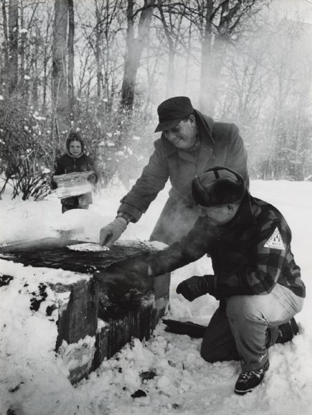 Two men cooking food wrapped in foil on a grate over a fire outdoors. One man's sleeve has a patch that reads: "Family Camping, MFCA Milwaukee." A child is walking toward them carrying a log. Snow is on the ground. Caption on back reads: "Camp Sidney Cohen on Upper Nemahbin Lake, 'family campout' sponsored by the Milwaukee Family Camping Association of the municipal recreation department Feb 1959."