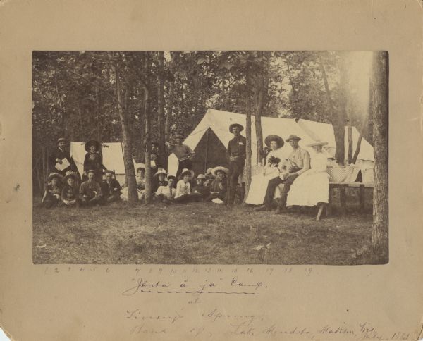 Nineteen men and women are posing outdoors in front of tents in a forest clearing. At the bottom the caption reads: "Jänta å ja" Camp at Livesey's Spring, Bank of Lake Mendota, Madison, Wis. July, 1885." The rear of the original photograph includes a list of people with their home towns: 1. Miss Belle Brown - Stevens Point, Wis.; 2. Mr. Chas. King - Madison, Wis.; 3. Miss Annie Stewart - Madison, Wis.; 4. Mrs. Chas. King - Madison, Wis.; 5. Howard B. Smith - Leon, Wis.; 6. George Main- Madison, Wis.; 7. Gene Hand - Racine, Wis.; 8. Virginia Ingman - Madison, Wis.; 9. May Brown - Steven's Point, Wis.; 10. George Waldo - Manitowoc, Wis.; 11. Will Cramer - Milwaukee, Wis.; 12. Kate McDonald - Trempeleau, Wis.; 13. George Bann - Madison, Wis.; 14. Fred Meyer - Lancaster, Wis.; 15. Ella Spaulding - Black River Falls, Wis.; 16. Fred Hooker - Milwaukee, Wis.; 17. Lizzie Hand - Racine, Wis.; 18. John Parkinson - Madison, Wis; 19. Mame Spaulding - Black River Falls, Wis. Miss May Newman, and I, Julius Ellison, were also members of the Camp, but left the day before this photograph was taken, i.e. July 3, 1886."