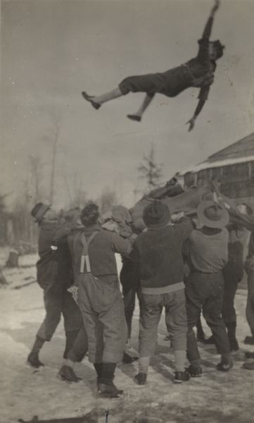 Group of lumberjacks entertaining themselves in camp by tossing a man in the air using a blanket. The man in the foreground with his back to the camera is identified as Leo.