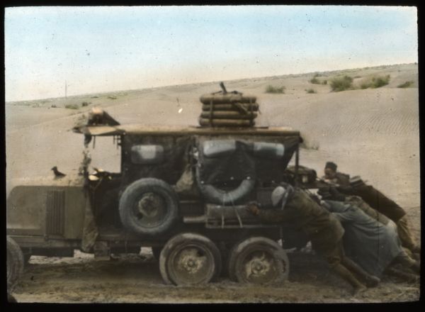 Hand-colored lantern slide showing a group of men on an archaeological expedition in Algeria pushing one of the expedition vehicles nicknamed "Hot Dog" in wet sand.