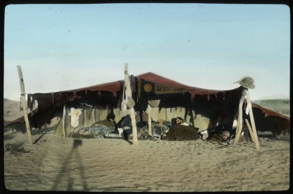 Members of the Algerian Expedition in the desert under the shelter of a tent open at the sides and front. The men are on the ground in sleeping bags. A hat is hanging on one of the wooden posts, and a Beloit College banner is hanging in the center. A shadow in the foreground may be from the camera on a tripod.