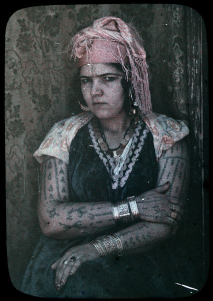 Autochrome portrait of a tattooed Tuareg woman posing in front of a patterned cloth hanging behind her. She is wearing a head wrap, as well as several bracelets, rings, earrings and necklaces.