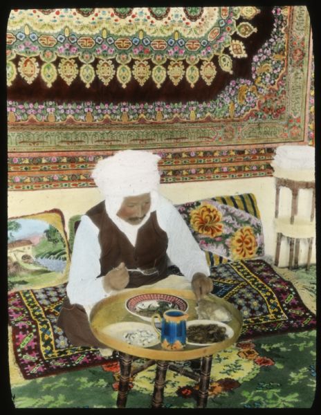 Hand-colored lantern slide showing an Caid Belaïd, Tuareg interpreter for Alonzo Pond's 1925 Algerian expedition, sitting on pillows and rugs on the floor and eating a meal in luxury. The room is filled with colorful tapestries.