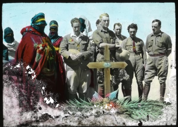 Hand-colored lantern slide of Alonzo Pond, Count Byron de Prorok and other members of the Algerian expedition paying respects to slain missionary Father Charles Eugène de Foucauld at El Ménia, Algeria. Veiled Tuareg men wearing colorful robes are standing on the left.