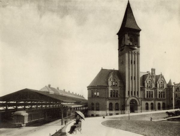 View of the Chicago & Northwestern Railroad station in Milwaukee. A couple is sitting on a bench under an umbrella in the foreground. The baggage car of a train is under the depot canopy at left.