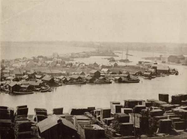 Elevated view of fishing village on Jones Island, Milwaukee. Several fishing boats are docked along the shoreline. Large piles of lumber are in the foreground across from the island.