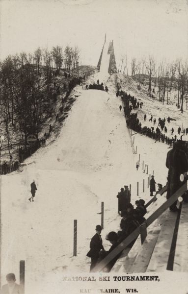 View from the bottom of a ski jump in Eau Claire, the host of a national ski tournament. Several people are standing along the sides of the landing area which is bordered by a fence. Other spectators are halfway up the hill, and wooden bleachers for more spectators are in the foreground. The area on both sides of the jump is wooded. Caption reads: "National Ski Tournament, Eau Claire, Wis."