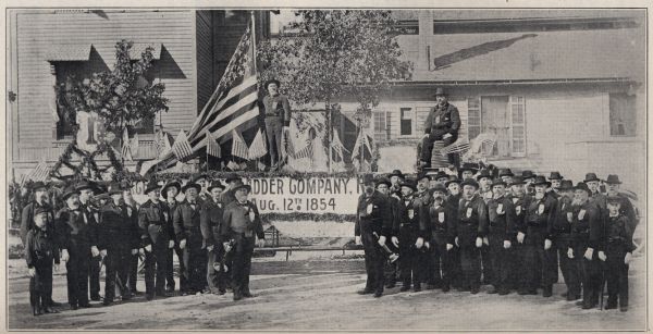 Outdoor group portrait of the members of Milwaukee Hook and Ladder Company No. 1. Two men are posing behind the group on a platform bearing several U.S. flags, and a banner, possibly recognizing the date the company was established, August 12, 1854.