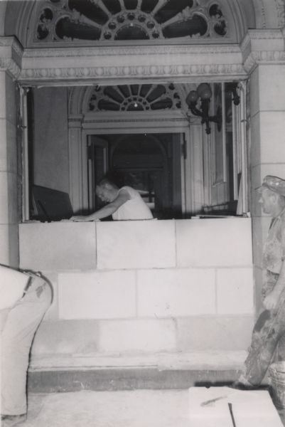 View of a worker laying concrete blocks to close off one of the three front entrances to the State Historical Society of Wisconsin headquarters building. Another worker is standing and watching from the right. This was part of a renovation project which saw first floor windows closed off to increase interior wall space.
