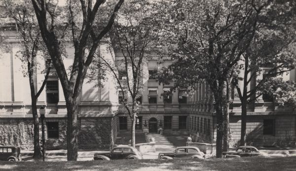 View from Bascom Hill and across Park Street towards the west facade of the State Historical Society of Wisconsin headquarters building, giving a view of the courtyard and west entrance. Vines are climbing up the walls of the building, and four cars are parked along the curb in front.