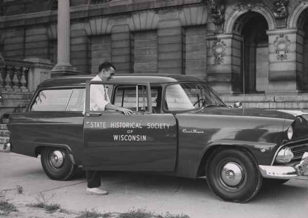 Don McNeil standing inside the open passenger door of the State Historical Society of Wisconsin's first fleet vehicle, a Ford Ranch Wagon. The vehicle is parked on Library Mall in front of the east entrance to the Society headquarters building.
