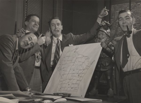 State Historical Society of Wisconsin staff enjoying a fun moment and posing with a map of Wisconsin and a gnome statue. Left to right: John Jacques, Clifford Lord, Harry Hunter, and Don McNeil.