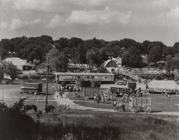 Elevated view from hill looking north, showing the grounds of the Circus World historic site, with visiting crowd. On display are several train cars and wagons, including Ringling Brothers Barnum & Bailey, Sells Forepaugh, Christy Brothers, and Miller Bothers. Many visitors are on a bridge spanning the Baraboo River, and a parking lot filled with cars is on the right. Barns that were used by the Ringling Brothers Circus and are now part of the Museum are across the river.