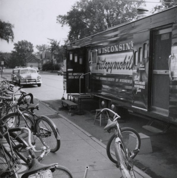 Several bicycles are parked on the sidewalk near the Wisconsin Historical Society Historymobile, parked near the curb. The Historymobile was visiting the Neville Public Museum.
