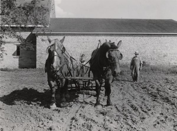 View from front towards two horses pulling a plow in a field being driven by a boy. On the right an older man is walking beside the team, and in the background is a stone building. The image is a still frame from a film of the Stonefield historic site for the movie <i>The Presence of our Past</i>.