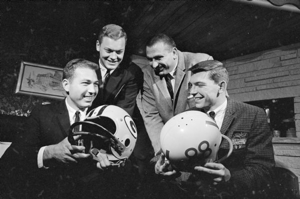 Bart Starr, quarterback, sitting front left, and Fred "Fuzzy" Thurston, guard, standing on the right, of the Green Bay Packers visiting with Jim Bakken, punter & place kicker, standing on the left, and Pat Richter, end, sitting in front on the right, of the Wisconsin Badger football team during a gathering at the Hoffmann House. Starr is holding a Packers helmet, and Richter is holding a Badgers helmet. Starr was in Madison to speak at the annual father and son banquet at Bethel Lutheran Church. 