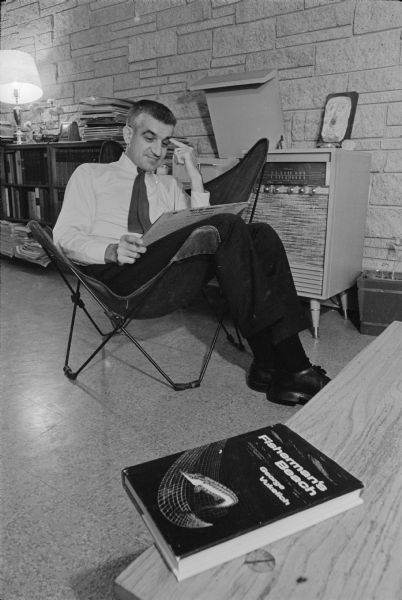 Radio host and newly published author, George "Papa Hambone" Vukelich relaxing at his home on Plymouth Circle. He is sitting in a chair holding an LP copy of Dick Ruedebusch's <i>Meet Mr. Trumpet!!!</i> which is playing on the stereo system in the background. A copy of his first novel, <i>Fisherman's Beach</i> is lying on a table in the foreground. A full bookshelf with a stack of record albums, a lamp, flowers, and a clock are in the background. 