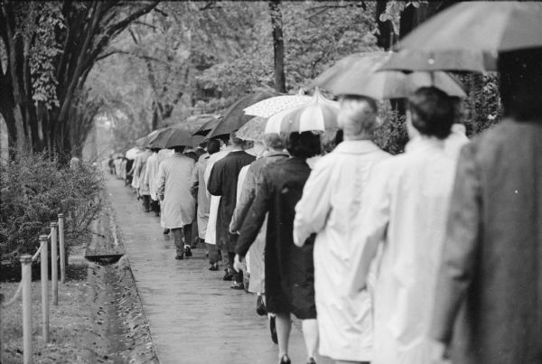 A long, orderly line of student sorority and fraternity members carrying umbrellas and marching in pairs to protest of the decision to ban the Delta Gamma sorority from the UW-Madison campus. Protestors walked up "Fraternity Row" on Langdon Street, up Bascom Hill, past Bascom Hall where they were met by Dean of Students, LeRoy Luberg, who was presented with a protest resolution by Robert Jennings, President of the Wisconsin Intrafraternity Association. The group of about 2,000 then walked back down the hill and back to Langdon Street. The decision to ban the sorority came after the national sorority suspended the Beloit chapter for pledging Patricia Hamilton, an African-American woman from Madison.