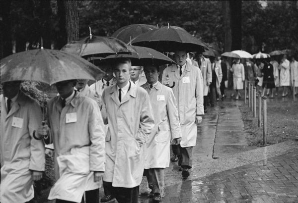 A long, orderly line of student sorority and fraternity members carrying umbrellas and marching in pairs to protest the decision to ban the Delta Gamma sorority from the UW-Madison campus. Protestors walked up "Fraternity Row" on Langdon Street, up Bascom Hill, past Bascom Hall where they were met by Dean of Students, LeRoy Luberg, who was presented with a protest resolution by Robert Jennings, President of the Wisconsin Intrafraternity Association. The group of about 2,000 then walked back down the hill and back to Langdon Street. The decision to ban the sorority came after national sorority suspended the Beloit chapter for pledging Patricia Hamilton, an African-American woman from Madison. The group is seen here nearing Bascom Hall with North Hall in the background.