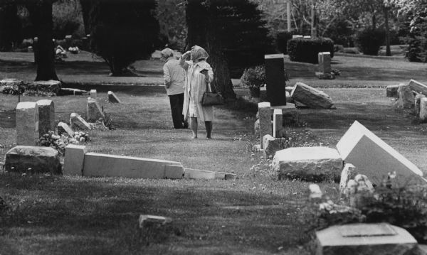 Two women are standing between rows of gravestones in a cemetery. Some of the gravestones have been knocked over. One woman is pointing over her shoulder while looking towards the left. Caption reads: "Tombstones that were pushed over by vandals were examined Sunday at the Holy Sepulcher Cemetery, 3800 E. College Ave., Cudahy."