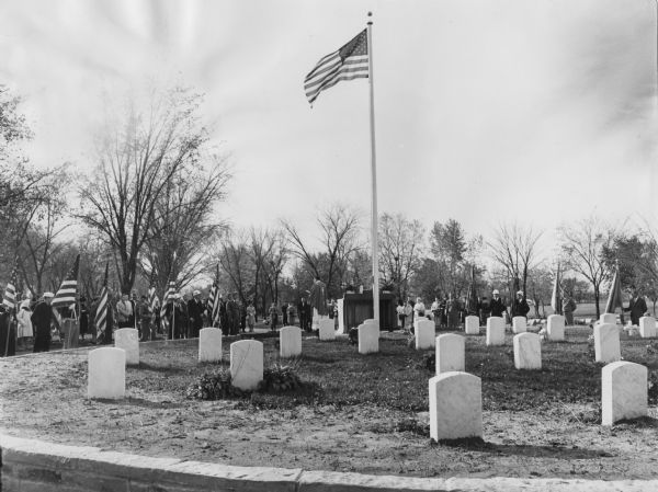 View towards a group of people standing in a cemetery around an altar and a flagpole with an American flag. A clergyman in an alb and chasuble standing in the center. A number of the assembled people in the group are wearing hard hats and military uniforms, with some of the men holding American flags. Caption reads: "Catholic War Vets will dedicate the Veterans Memorial mound in Holy Sepulcre (<i>sic</i>) Cemetery, W. College oa. (<i>sic</i>) and S. Lake Dr. Cudahy on Sunday. American Legion, Polish Legion and V.F.W. will participate in the ceremonies there at 9:00 AM. Sunday, 200 CT. Guess it will be quite a local turnout. Gonyea will make the principal address and four firing squads will rend the air..." 