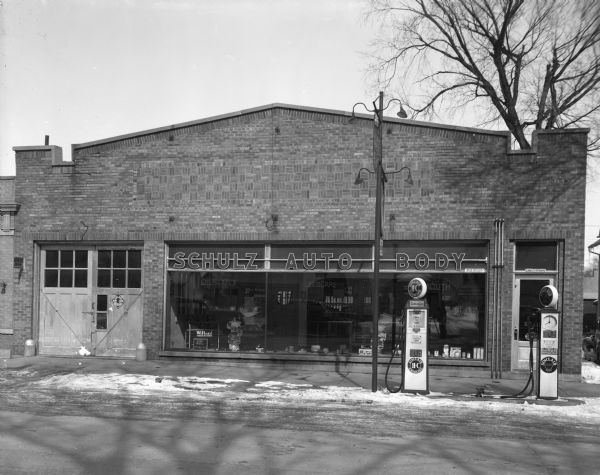 View of the exterior of the Schulz Garage. Two gas pumps are at the curb. Large, plate glass windows are reflecting automobiles parked across the street, and the photographer.