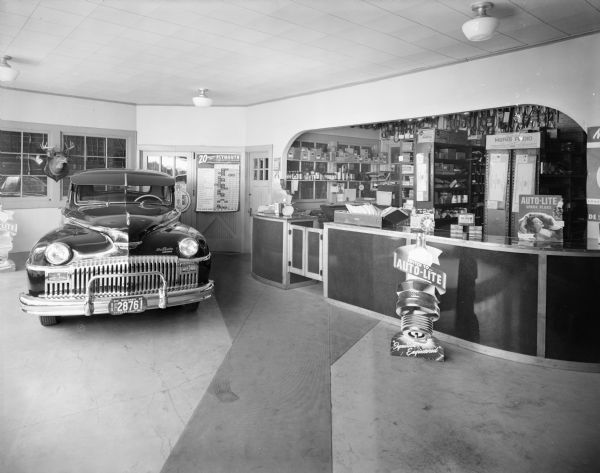 A DeSoto Fluid Drive automobile is parked on the left, and has a Wisconsin license plate mounted on the front with a date of 1946. On the right is a service counter, and behind it are shelves of parts. A spark plug advertisement for "Auto-Lite" is set up on the floor in front of the counter.