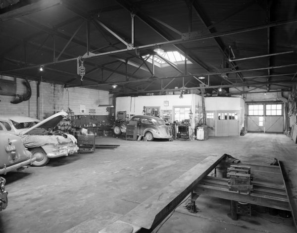 Interior view of the garage. In the foreground is a lift, and in the background on the left is the office, which is under a skylight. Garage doors are to the right of the office. Cars are parked on the left, and one has the hood lifted and tarps have been places around the engine. Another car is near the office, and the front is raised up on ramps. Tools and workbenches are along the wall and on carts, and parts are hanging on the walls.