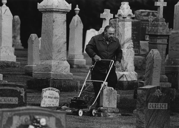 A man is using a lawn mower between rows of gravestones. He has turned from it and is looking at the ground. Caption reads: "CEMETERY TRIM - Roger Reif, of Beechwood in Sheboygan County, paused to check his work while mowing the grass at St. John's Lutheran Church Cemetery. Reif is the caretaker of the cemetery, on County Highway S just outside Beechwood."