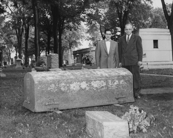 Two men in suits are standing in a cemetery, looking at a metal grave vault. The vault is supported by two wooden beams. Caption reads: "A 12 gauge Clark metal vault after 27 years underground in Riverside Cemetery, Oshkosh. Vault in perfect condition after being hoisted up end-wise by chain. Original paint intact."