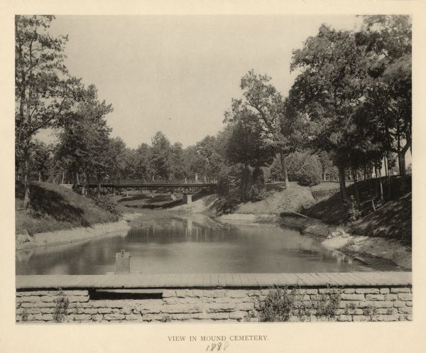 View towards a wooden walkway over a stone wall, perhaps a spillway, crossing a body of water. A bridge is in the background.