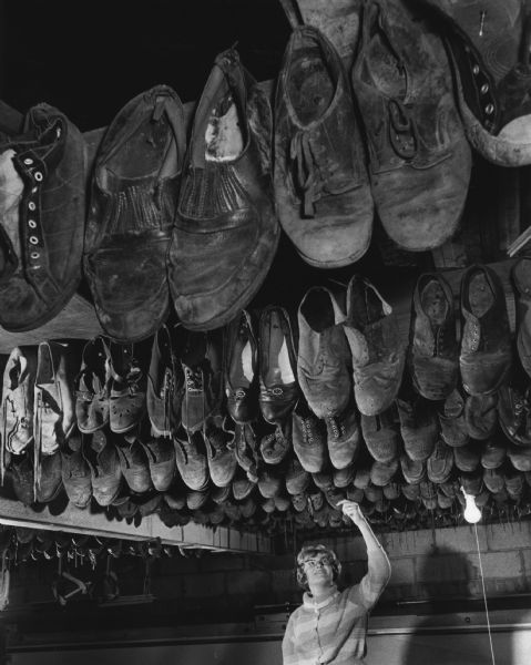 A woman is standing under rafters in a garage on which many shoes have been affixed. She is reaching up and touching the toe of one shoe. Caption reads: "For another three years, Barbara Kojis' shoes will be added by her father, George A. Kojis, 51, of 2208 S. 63rd St., West Allis, to the collection she viewed. He has nailed shoes of his other three children to the rafters in his garage from their first baby shoes until they reached the age of 16. Barbara is 13."